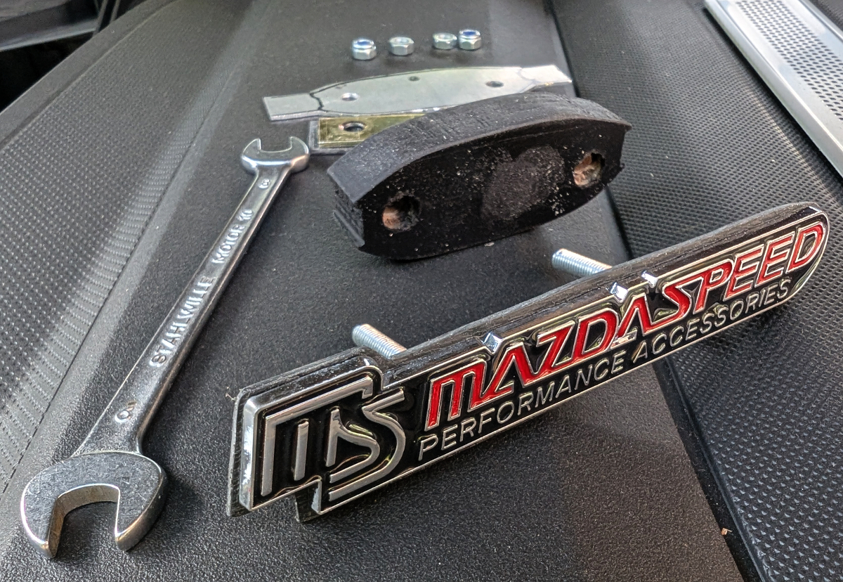 Emblem "Mazdaspeed" für den Kühlergrill; gibt es in der Bucht. Zusätzlich selbstgebauter Lückenfüller aus einer Siebdruckplatte passgenau für "in die Raute" (schwarz), ein Alublech mit etwas größer als die Raute, Befestigungsplatte, Schrauben.